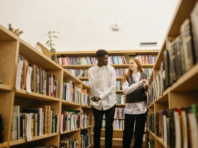 A black man and caucasian woman discussing in a library aisle surrounded by bookshelves.